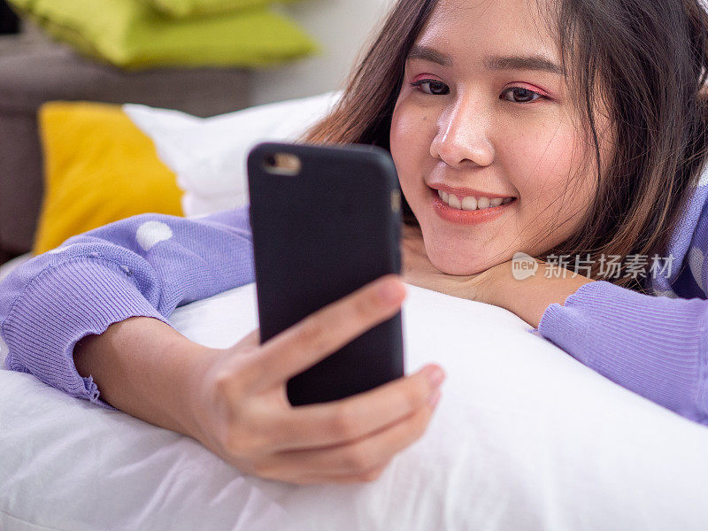 An Asian woman is lying on the bed, using the phone with a bright smile. Women are happy to use the phone and watch the series on mobile phones. Relax and enjoy concept